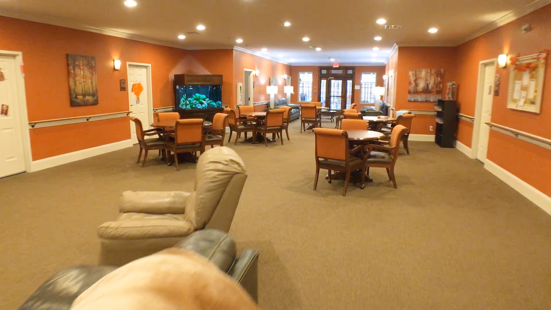 A spacious common area in a senior living facility with multiple round tables and chairs arranged throughout the room. There are comfortable armchairs in the foreground, warm orange walls with framed artwork, and a large fish tank against one wall. The room is well-lit with ceiling lights and lamps, and there are double glass doors at the far end.
