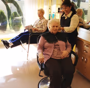 An elderly woman sitting in a salon chair with a black cape around her shoulders, receiving a hair treatment from a smiling hairdresser. In the background, another elderly woman is seated near a window, reading a magazine.