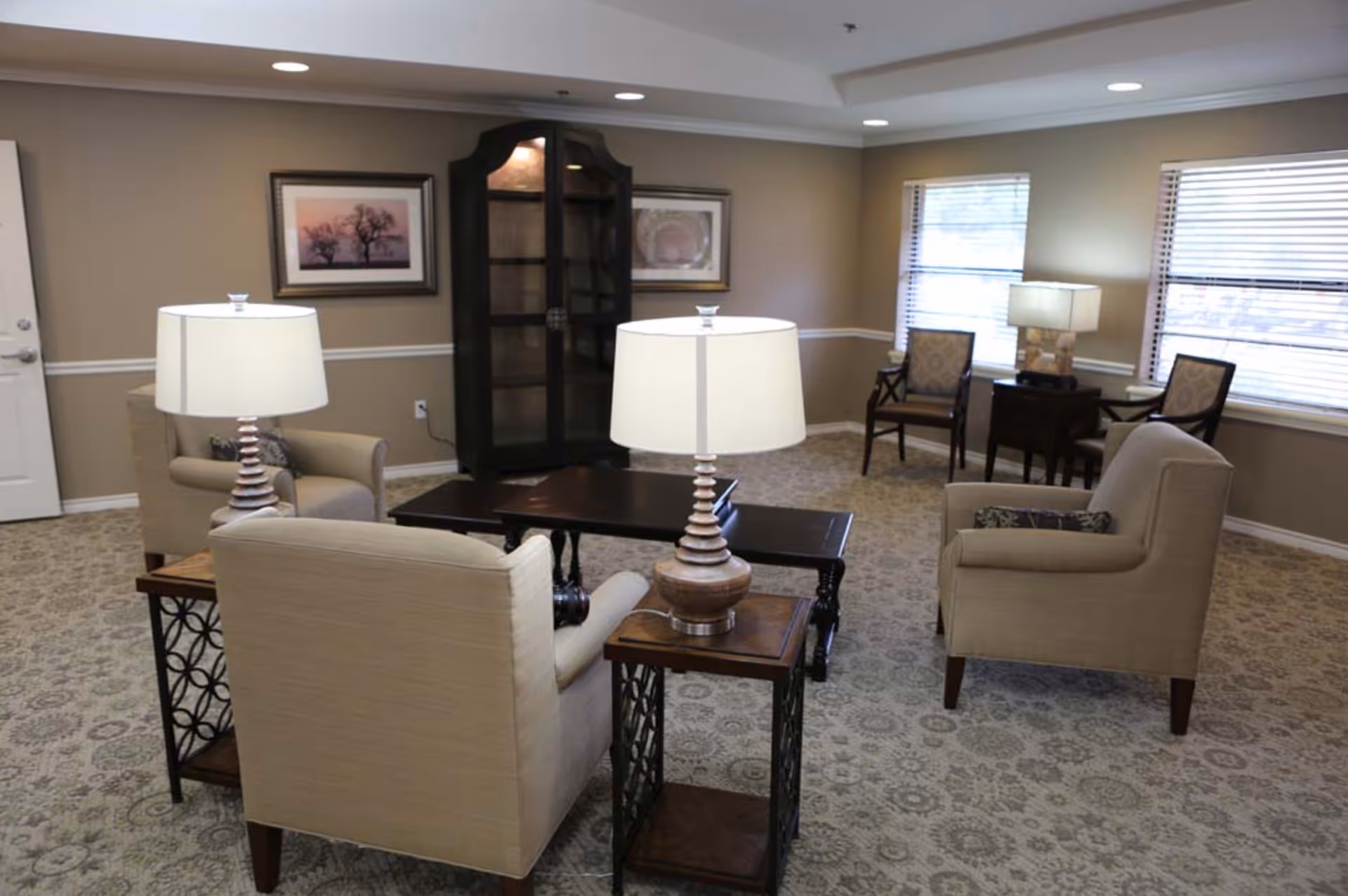 A cozy living room area with beige armchairs, a dark wooden coffee table, side tables with lamps, patterned carpet, two framed pictures on the wall, and windows with blinds letting in natural light.