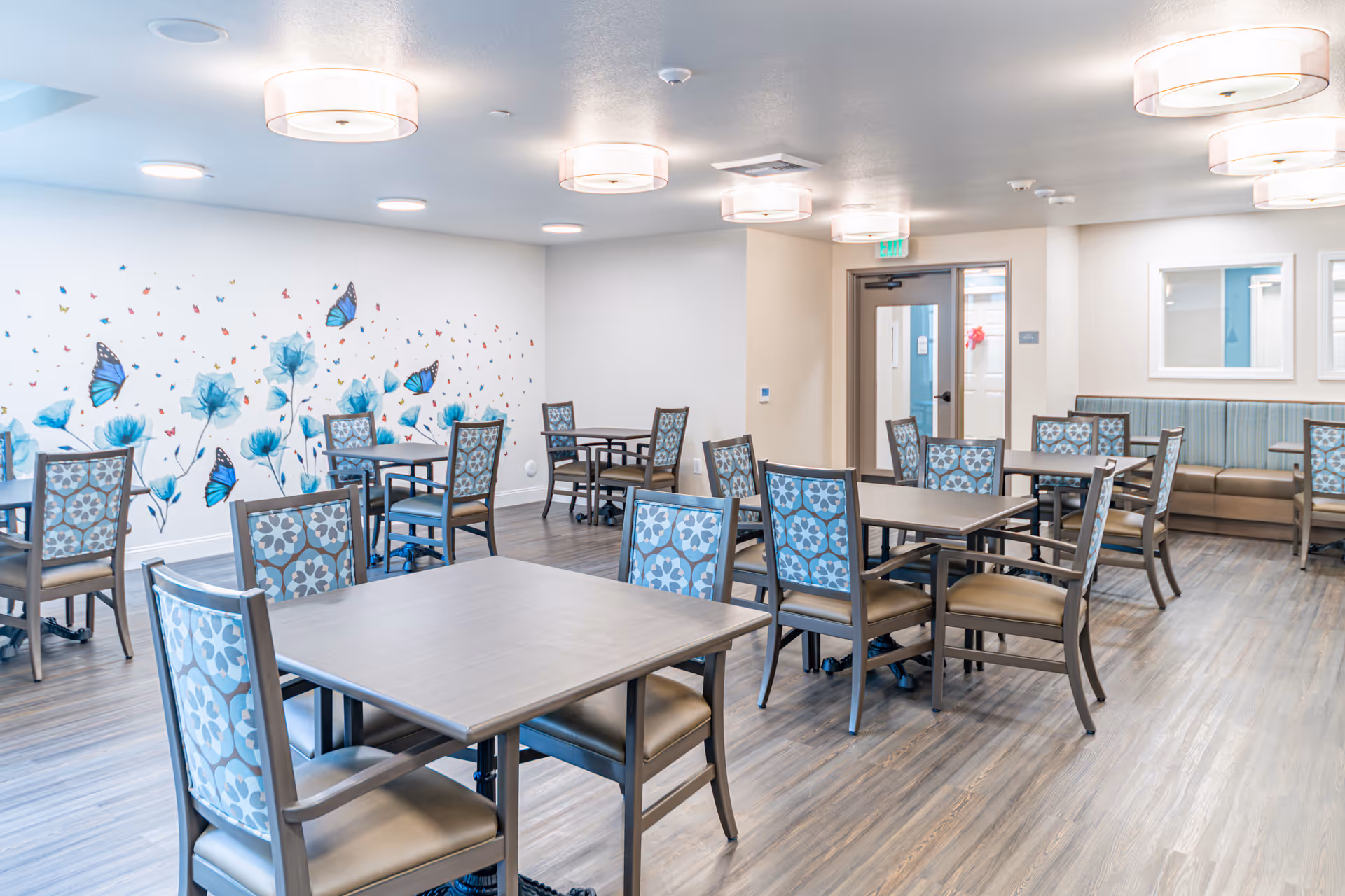 A bright and clean dining room in a senior living facility with multiple tables and chairs arranged neatly. The chairs have patterned blue upholstery, and the walls are decorated with a mural of blue flowers and butterflies. The room has modern ceiling lights and wood-style flooring.