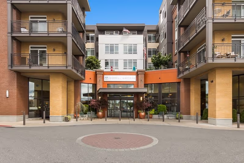 Front exterior view of Merrill Gardens at Kirkland, showing the entrance with glass doors, potted plants, and balconies on the upper floors of the building.