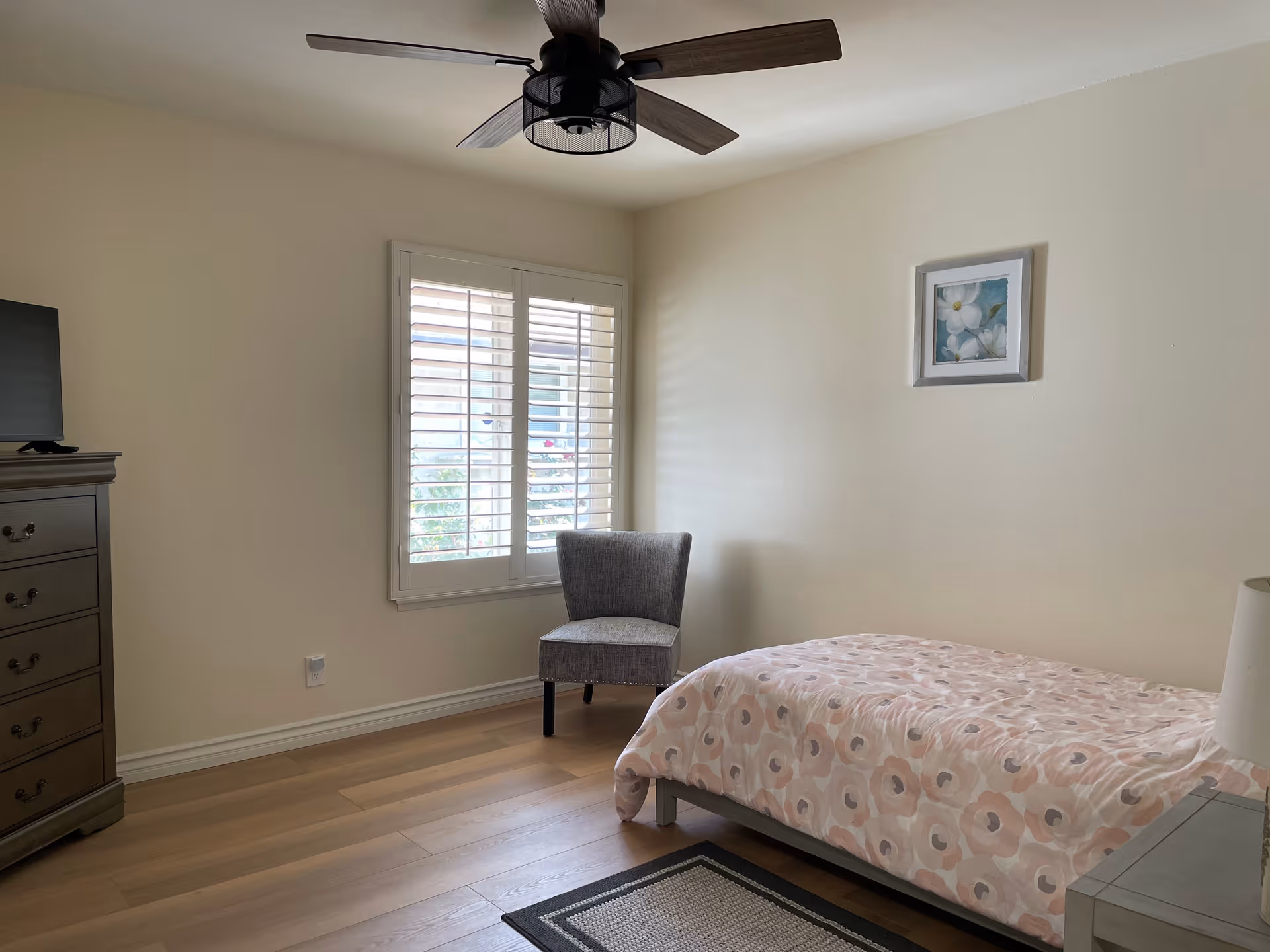 A simple bedroom with a single bed covered in a pink floral patterned comforter, a gray upholstered chair near a window with white shutters, a wooden dresser with a TV on top, a ceiling fan with wooden blades, and a framed floral picture on the wall.