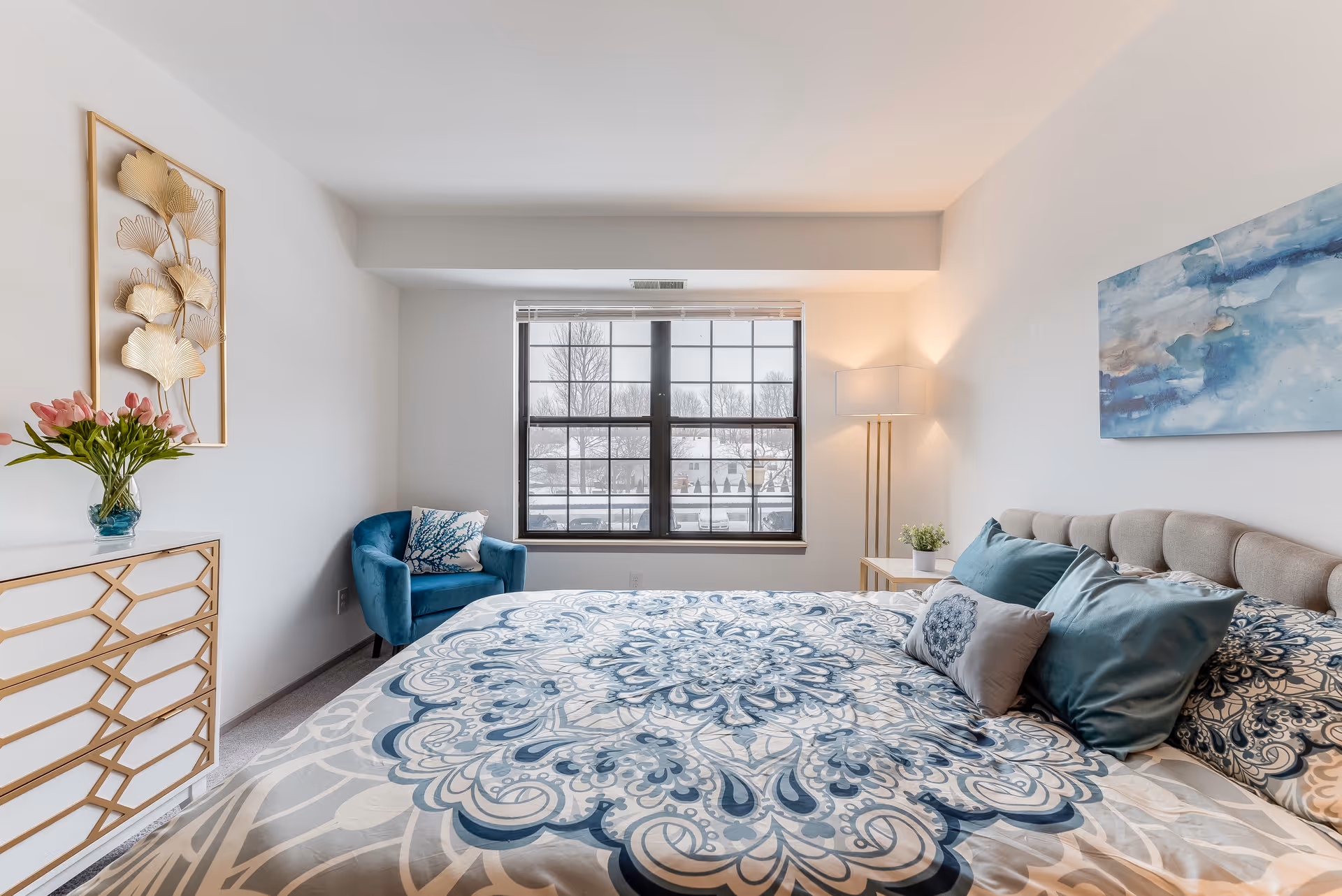 A bright bedroom with a large bed covered in a blue and beige patterned comforter and multiple pillows. There is a blue armchair with a decorative pillow near a window with a snowy outdoor view. A white dresser with gold accents and a vase of pink flowers is on the left side. The walls are white with a gold leaf wall decoration and a blue abstract painting. A floor lamp and a small side table with a plant are next to the bed.