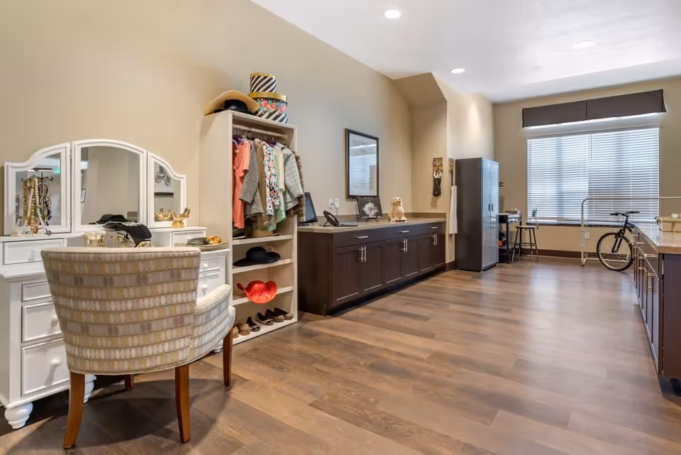 A spacious room with wooden flooring featuring a white vanity table with a mirror and jewelry, a beige patterned chair, an open wardrobe with hanging clothes and hats, a long dark wooden cabinet with a mirror and decorative items on top, a tall gray cabinet, a desk with a chair near a large window with blinds, and a bicycle parked by the window.