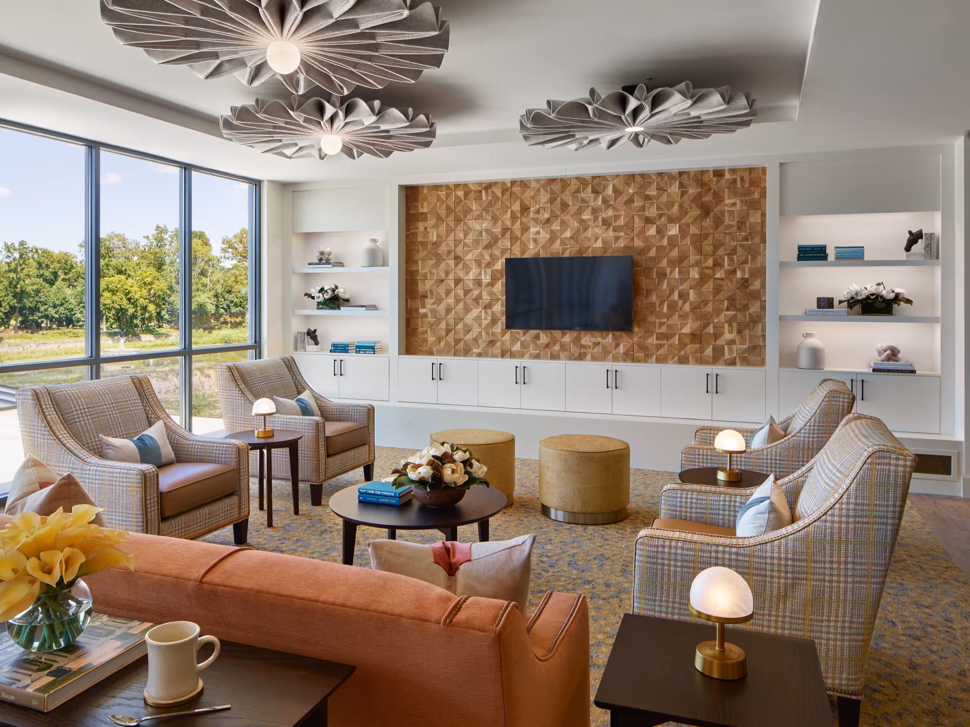 A bright and modern living room with large floor-to-ceiling windows showing greenery outside. The room features a peach-colored sofa, four plaid armchairs arranged around a round coffee table with a flower arrangement and books, two round ottomans, built-in white shelves with decorative items, and a wall-mounted flat-screen TV on a textured wooden accent wall. Unique ceiling light fixtures hang above.