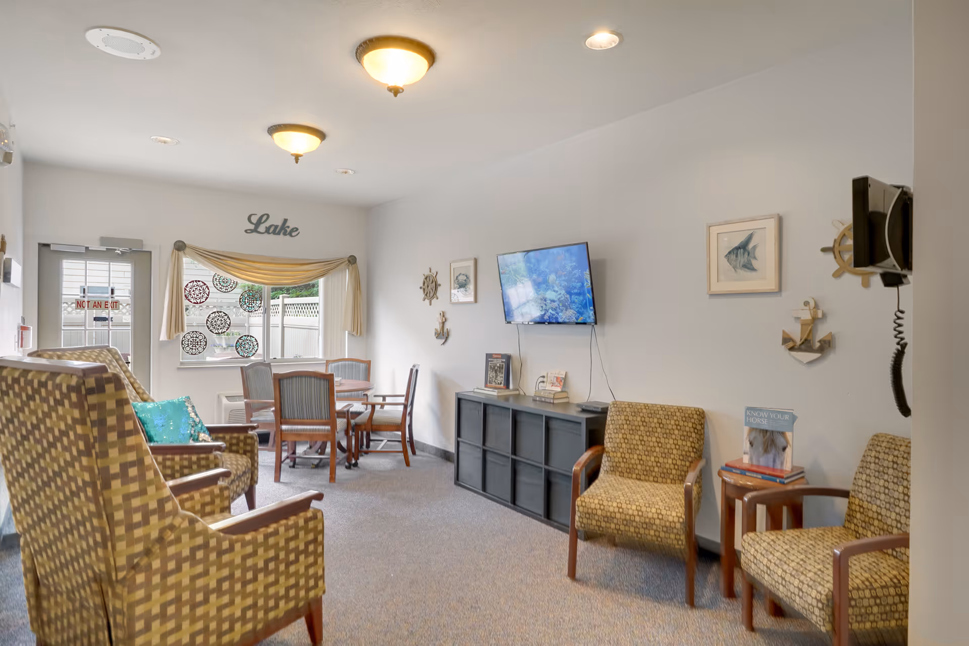 A cozy common area with patterned armchairs, a small table with chairs near a window, a wall-mounted TV, and nautical-themed decorations including a ship wheel, anchor, and fish artwork. The word 'Lake' is displayed above the window.