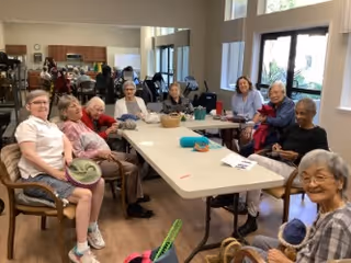 A group of elderly people sitting around a long table in a bright room with large windows, engaging in a social or craft activity. The room has wooden flooring and light-colored walls, with some kitchen cabinets visible in the background.