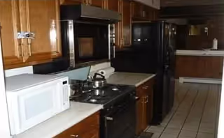 Interior view of a kitchen featuring wooden cabinets, a white microwave, a black stove with a kettle on it, a black refrigerator, and tiled flooring.
