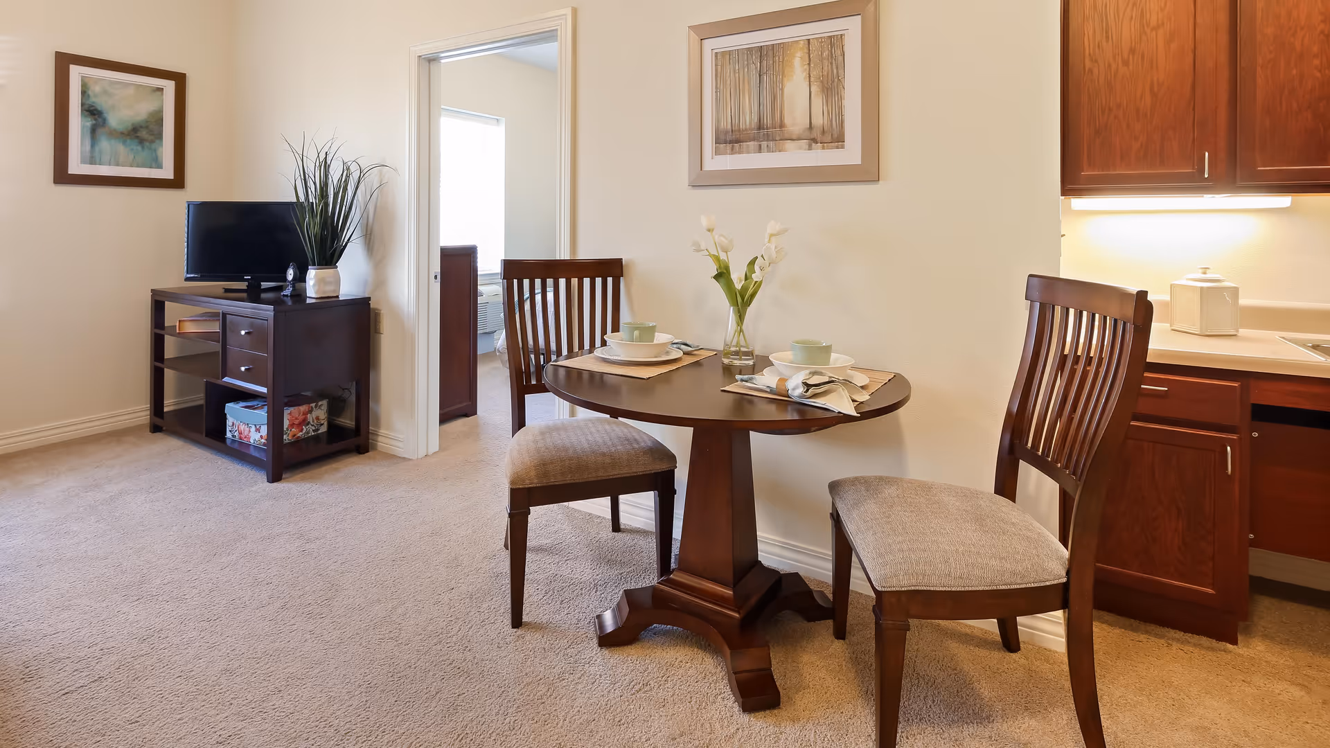 A cozy interior space featuring a small round wooden dining table set for two with dishes and a vase of flowers. Two wooden chairs with cushioned seats are placed around the table. To the right, there is a kitchen area with wooden cabinets and a countertop. On the left side, a dark wooden TV stand holds a small television and a decorative plant. A doorway leads to another room with a bed visible. The walls are light-colored and decorated with framed artwork.