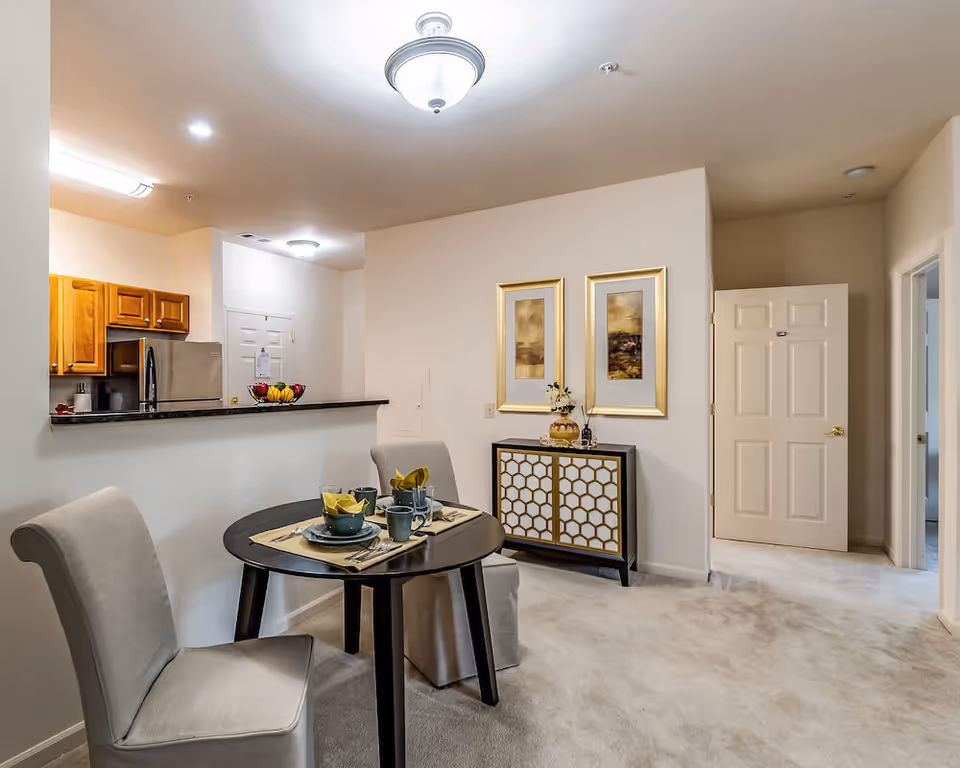 Interior view of a senior living facility dining area with a small round table set for two with dishes and napkins. Two upholstered chairs are placed around the table. In the background, there is a kitchen with wooden cabinets and a stainless steel refrigerator. A decorative cabinet with two framed artworks above it is against the wall. The room is carpeted and well-lit with ceiling lights.