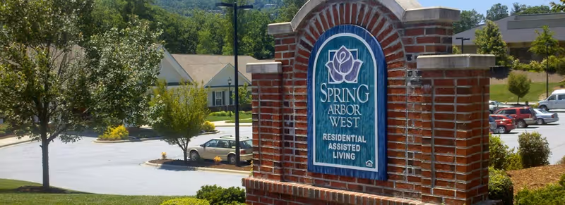 Brick entrance sign reading "Spring Arbor West Residential Assisted Living" with a parking lot, trees, and buildings in the background.