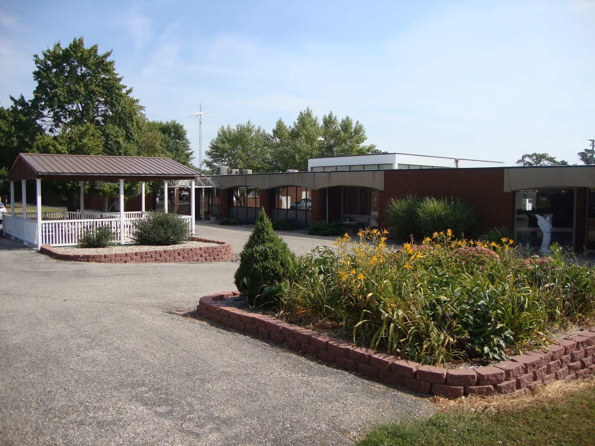 Exterior view of Goldwater Care Roseville facility showing a single-story brick building with large windows, a covered entrance area, and landscaped garden beds with green shrubs and yellow flowers. There is a paved driveway and a small gazebo structure with a brown roof and white railing on the left side.