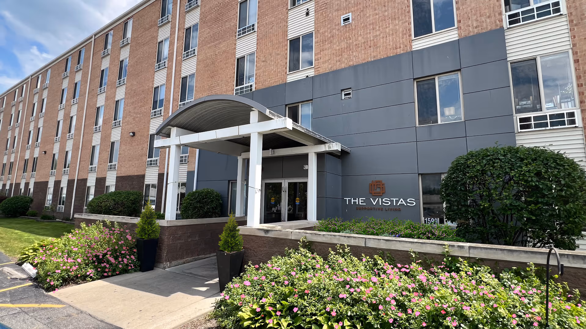 Exterior view of The Vistas of Fox Valley building entrance with a covered walkway, surrounded by green bushes and pink flowers under a partly cloudy sky.