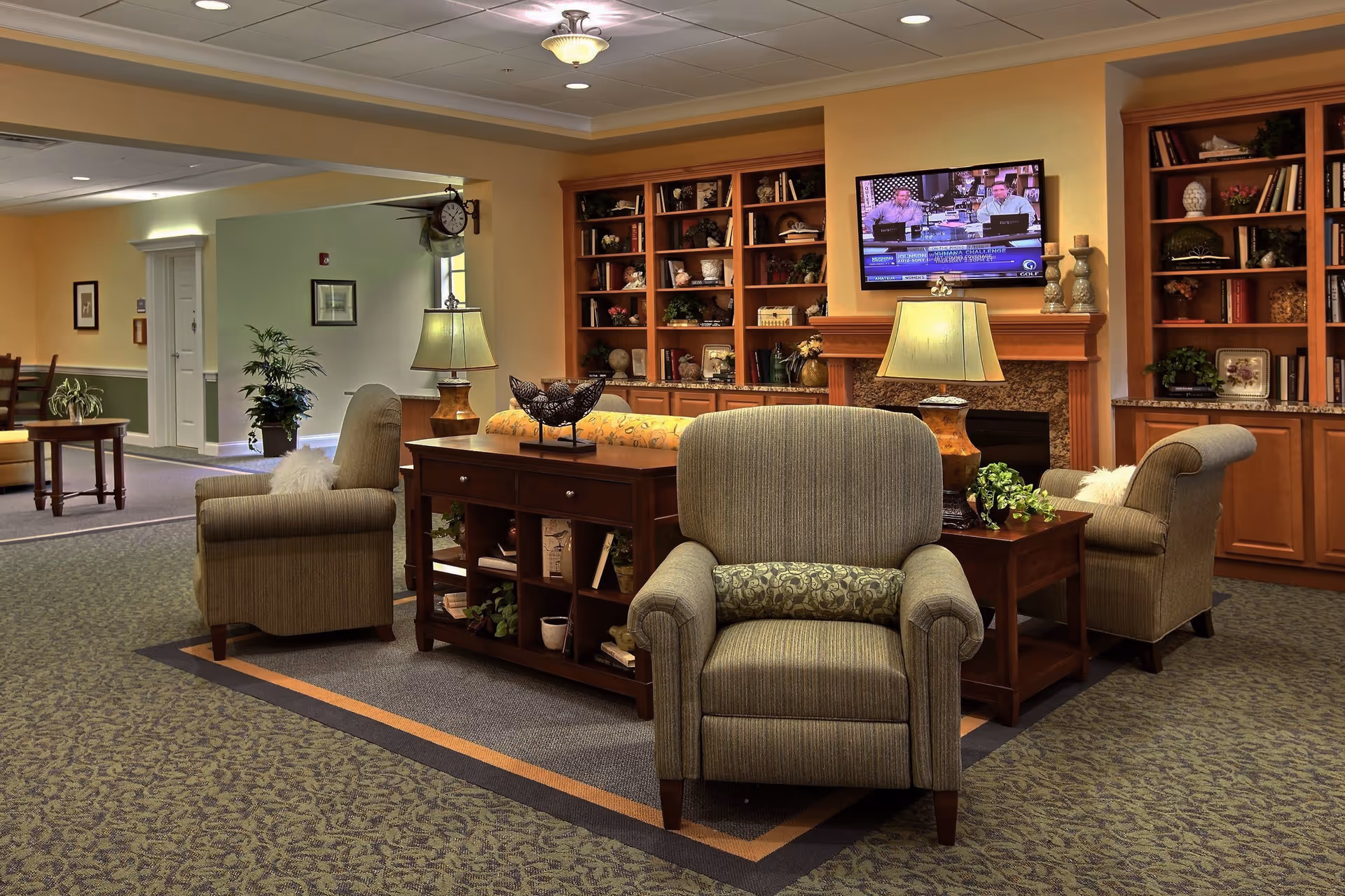 A cozy living room area in a senior living facility with several upholstered armchairs arranged around a wooden console table. The room features built-in wooden bookshelves filled with decorative items and books, a fireplace with a mounted flat-screen TV above it, and two table lamps providing warm lighting. The carpeted floor and soft yellow walls create a welcoming atmosphere.