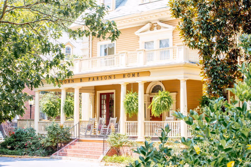 Front exterior view of the John B. Parsons Home, a large yellow building with white columns and a porch featuring rocking chairs and hanging plants, surrounded by greenery and trees.