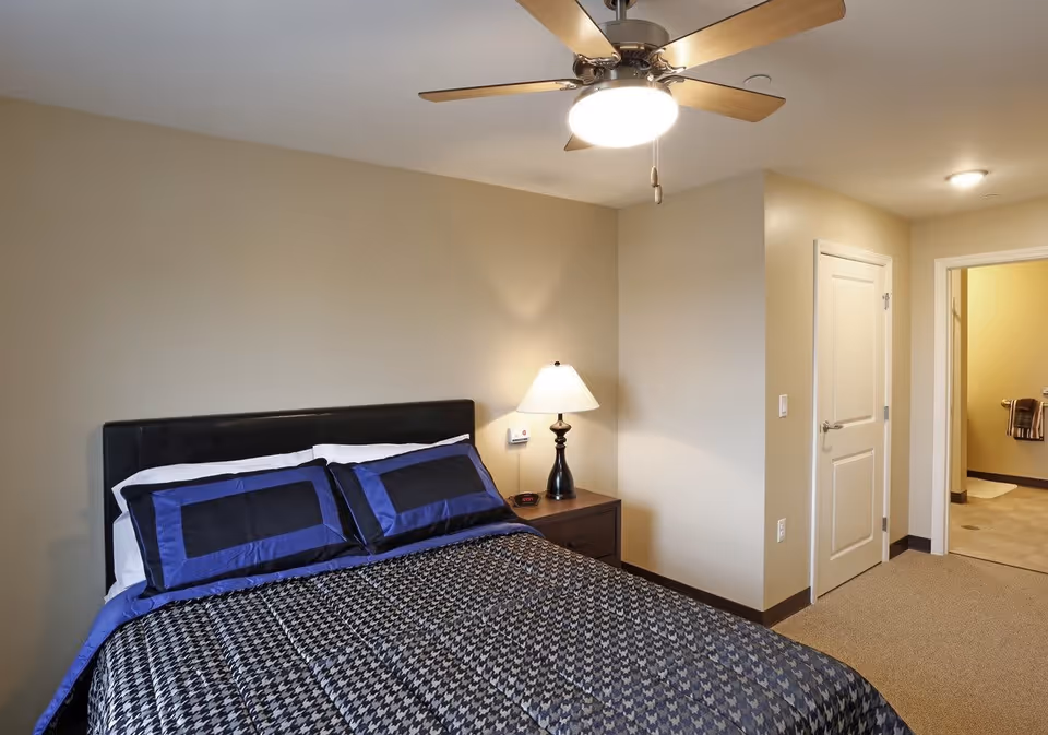 A bedroom with a bed featuring a black and blue patterned comforter and matching pillows. There is a nightstand with a lamp and an alarm clock beside the bed. The room has beige walls, a ceiling fan with a light, and a doorway leading to a bathroom.