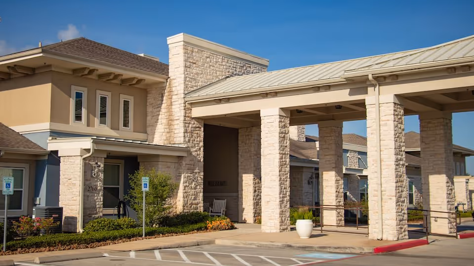 Exterior view of Orchard Park at Victory Lakes Assisted & Senior Living facility showing a stone and stucco building with a covered entrance supported by stone pillars, handicap parking spaces, and landscaping under a clear blue sky.