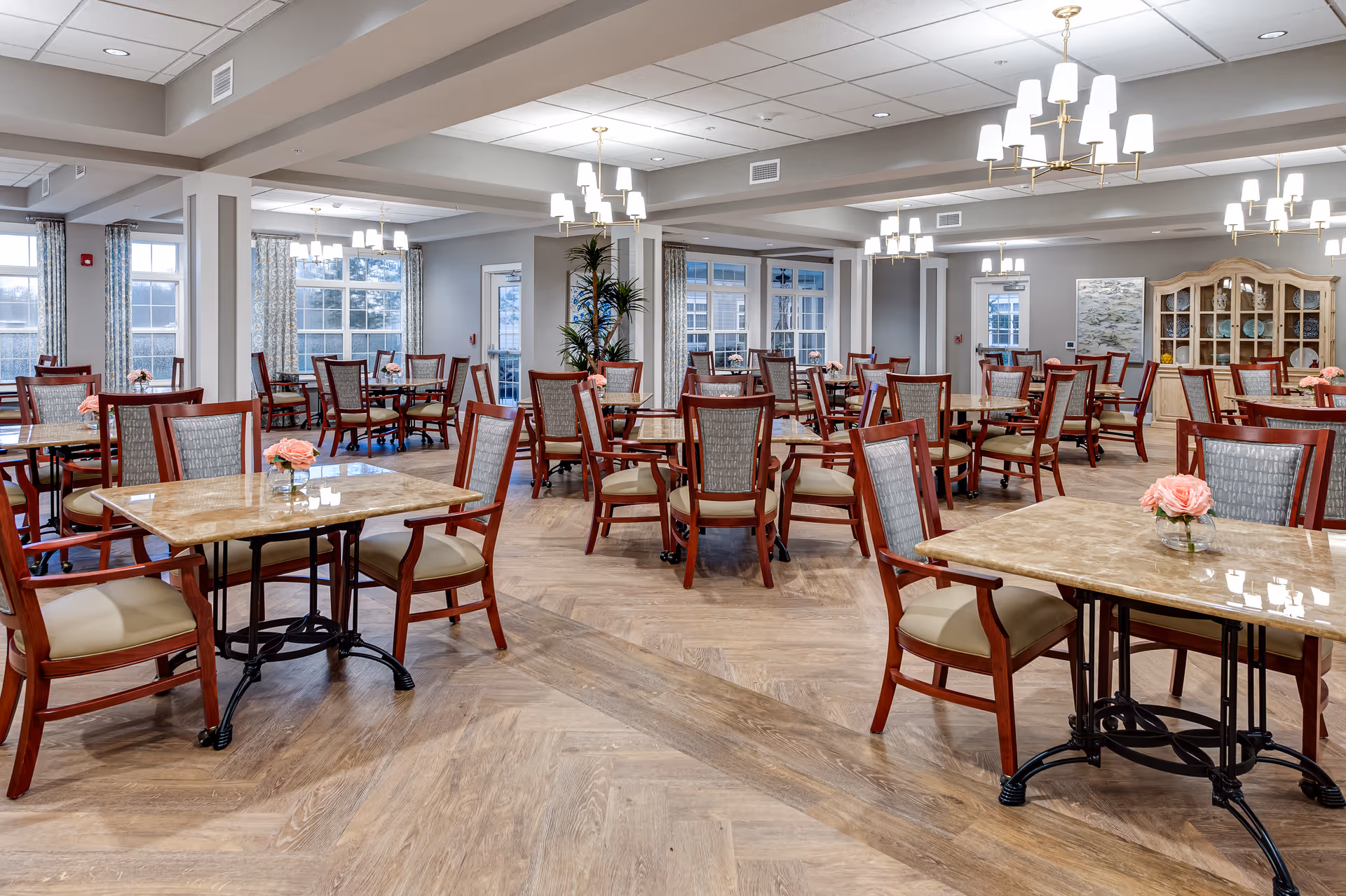A spacious dining room in a senior living facility with multiple tables and chairs arranged neatly. Each table has a small vase with pink flowers. The room features large windows with patterned curtains, wooden flooring, and several modern chandeliers providing warm lighting. A wooden cabinet with dishes is visible in the background.