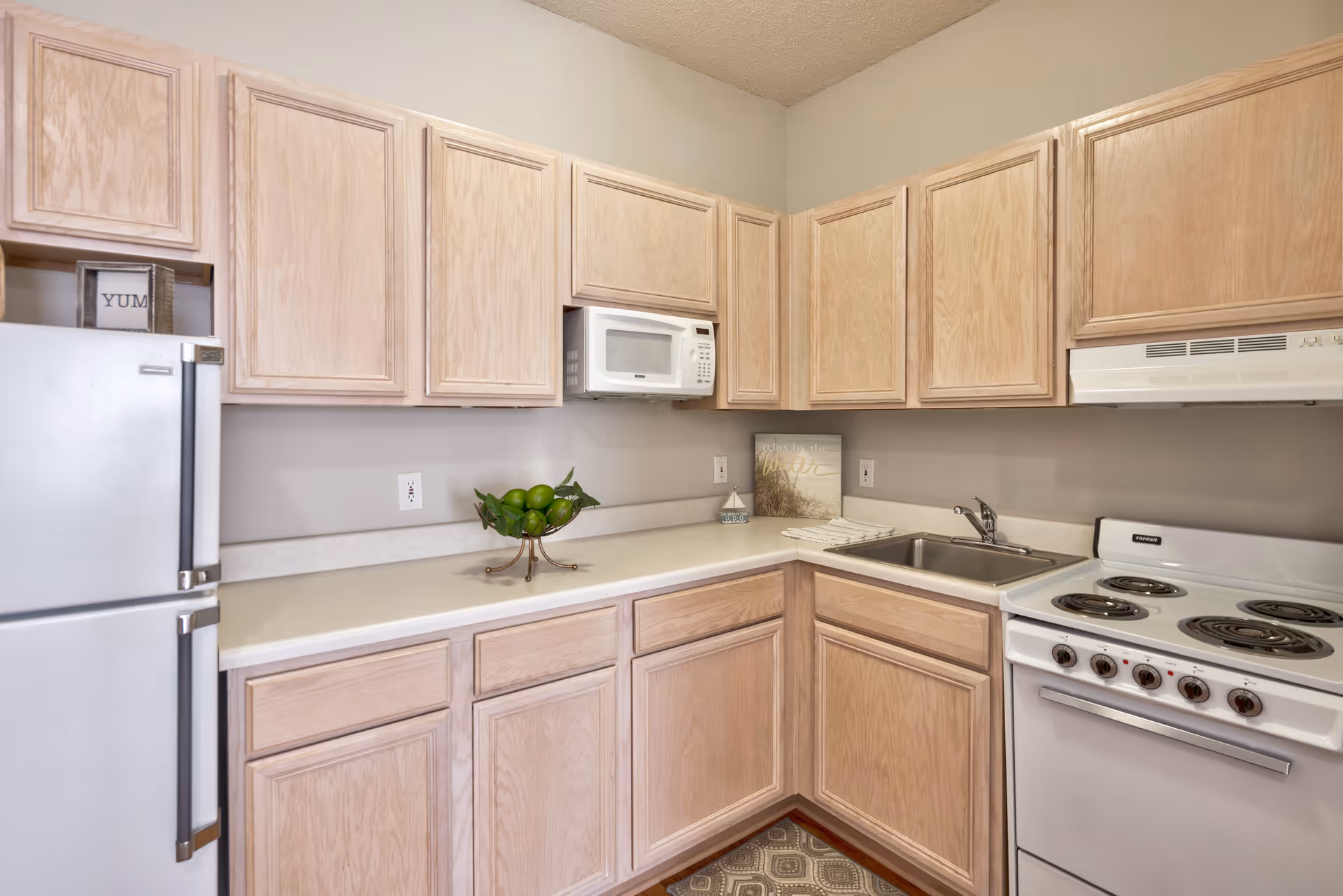 A compact kitchen with light wood cabinets, a white refrigerator, a white microwave mounted above the counter, a stainless steel sink, and a white electric stove with four burners. The countertop is light-colored and has a decorative bowl with green apples and a small framed sign that says 'YUM'.