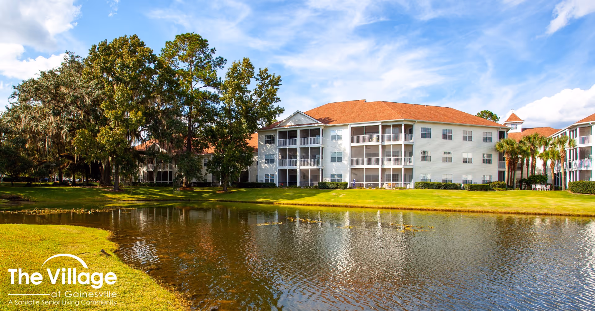 Exterior view of The Village at Gainesville senior living community showing a large white multi-story building with screened balconies, surrounded by green grass, trees, and a pond in the foreground under a partly cloudy blue sky.