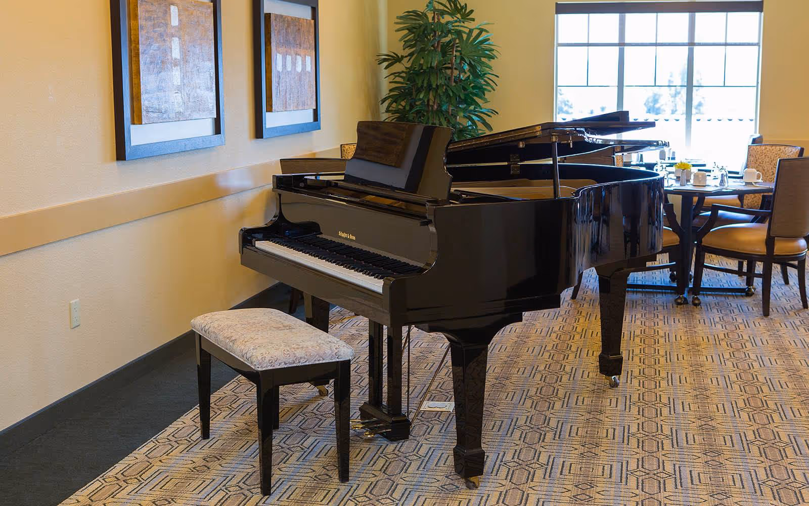 A black grand piano with a bench in a bright communal room with a dining table, chairs, and framed artwork on the wall.