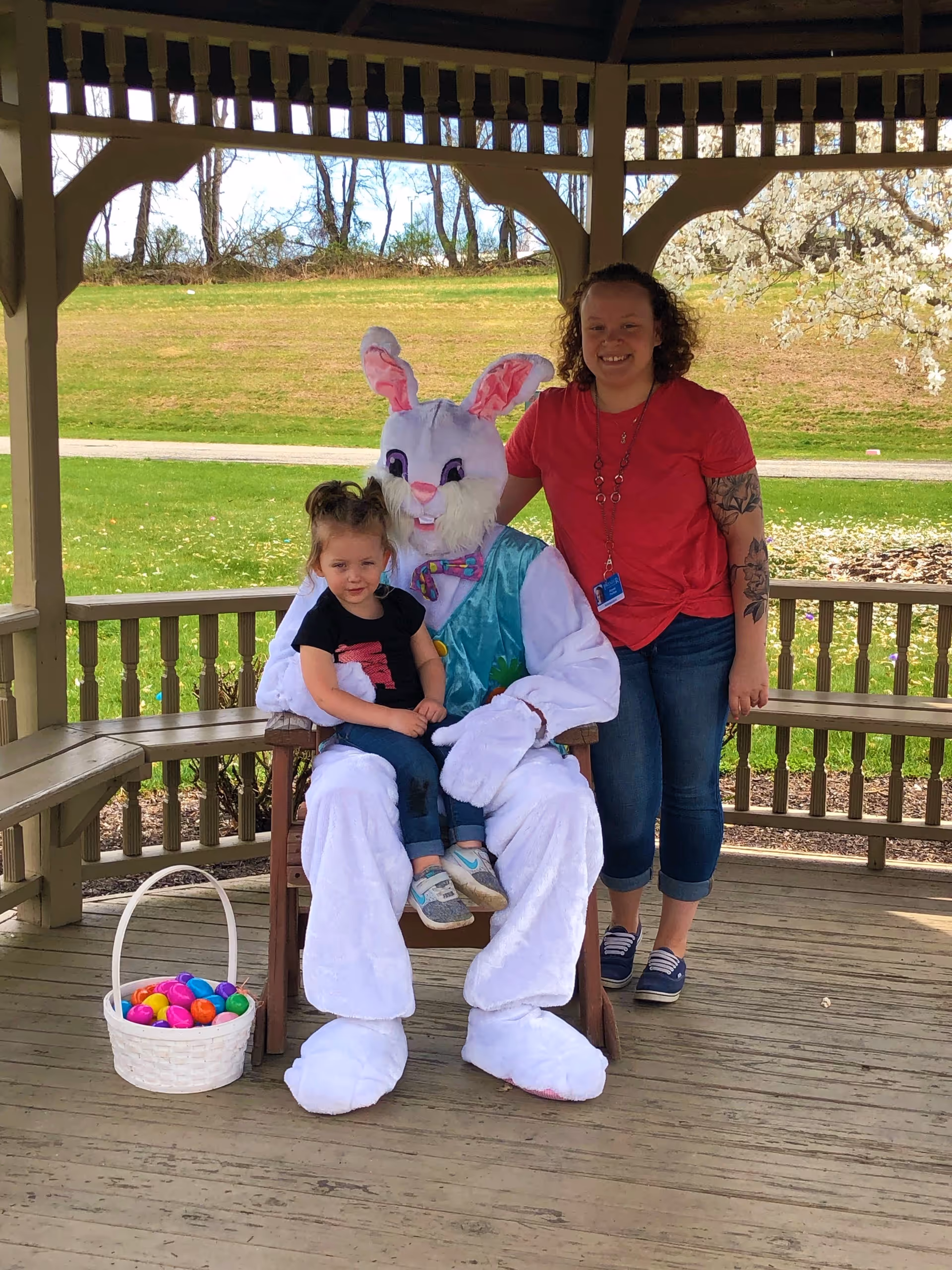 A young girl sitting on the lap of a person dressed in a white Easter Bunny costume with a colorful vest and bow tie, seated on a wooden chair under a gazebo. A woman in a red shirt and jeans stands next to them, smiling. A white basket filled with colorful plastic Easter eggs is placed on the wooden floor nearby. The background shows a grassy area with trees and a blooming tree with white flowers.