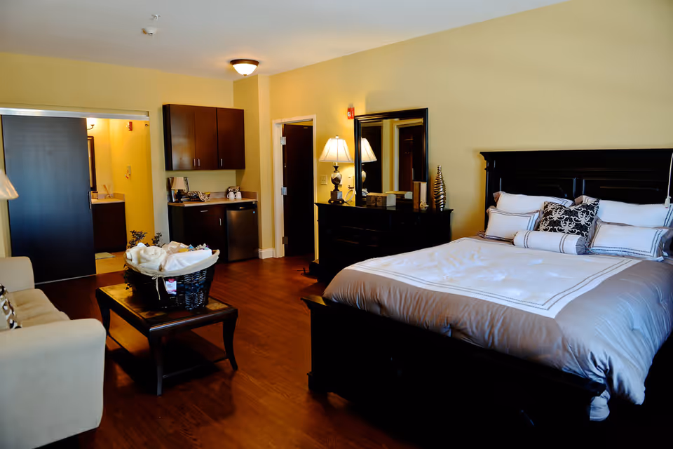 A spacious senior living bedroom with a large bed featuring multiple pillows and a gray and white comforter. There is a dark wooden dresser with a mirror and a lamp on top. A beige sofa and a wooden coffee table with a basket of towels are also visible. The room has wooden flooring and light yellow walls. In the background, there is a small kitchenette area with cabinets, a mini fridge, and a sink, as well as a doorway leading to a bathroom.