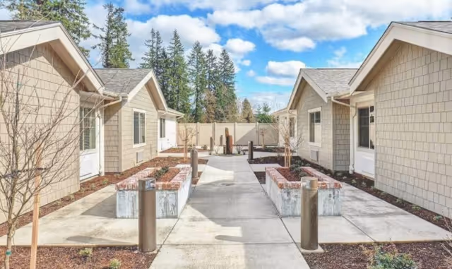 Courtyard between single-story memory-care cottages with concrete walkways, raised planter boxes and small trees under a blue sky.