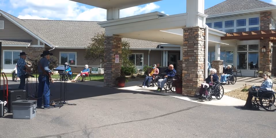 Residents in wheelchairs seated outside the covered entrance of a senior living facility while musicians perform nearby.