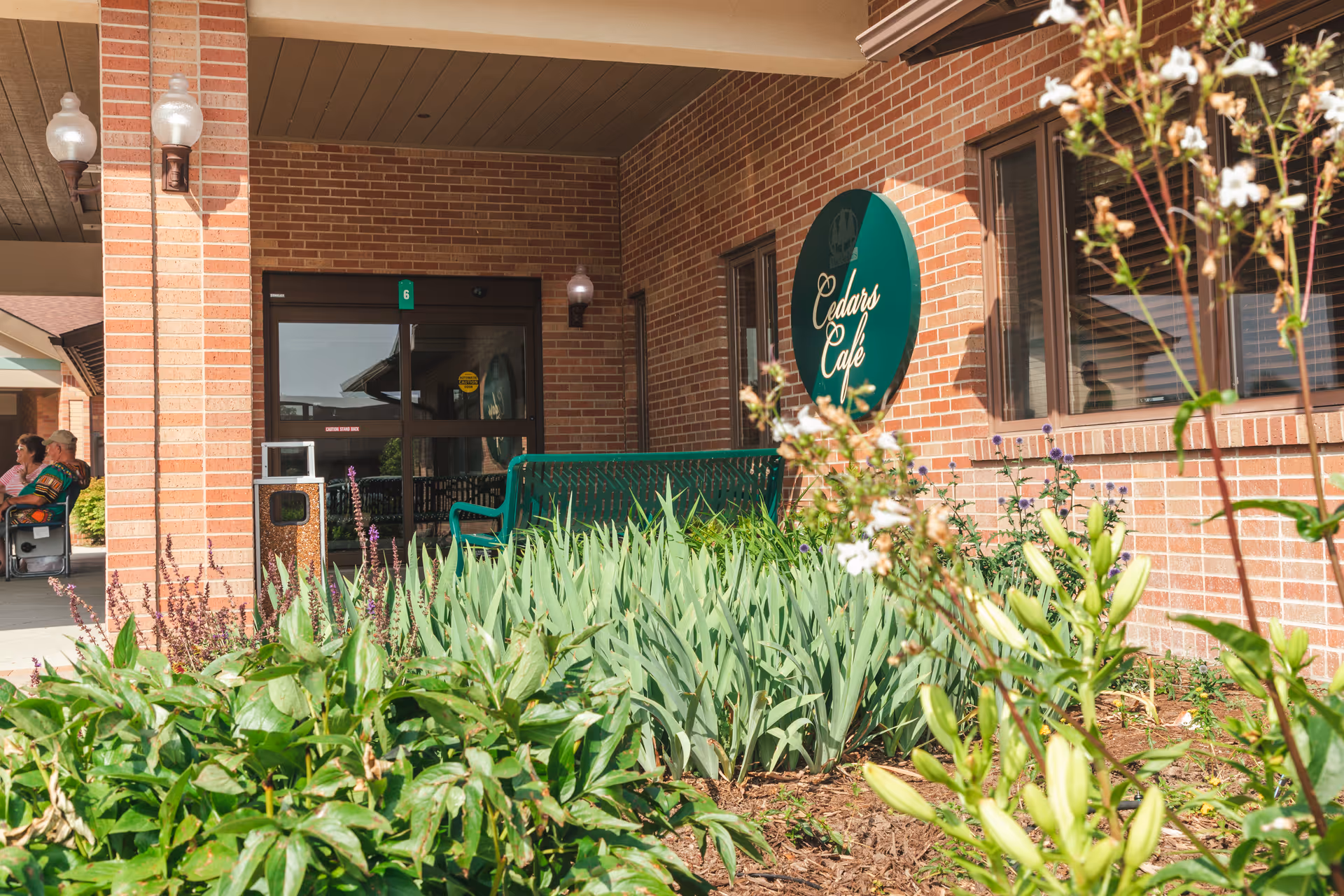 Outdoor view of a brick building with a green sign that reads 'Cedars Cafe'. There is a green bench near the entrance, surrounded by plants and flowers. Two people are sitting on chairs under a covered walkway to the left.