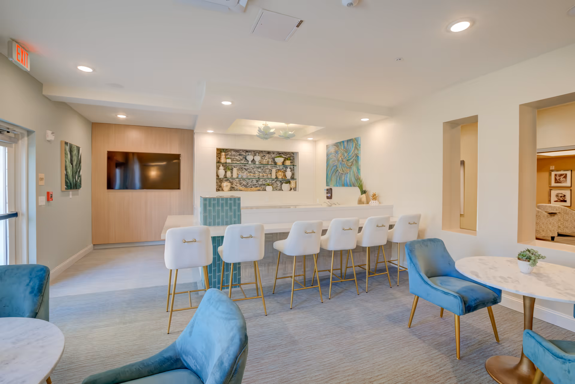Bright communal lounge with a long white bar, six white stools, teal chairs and small round tables.