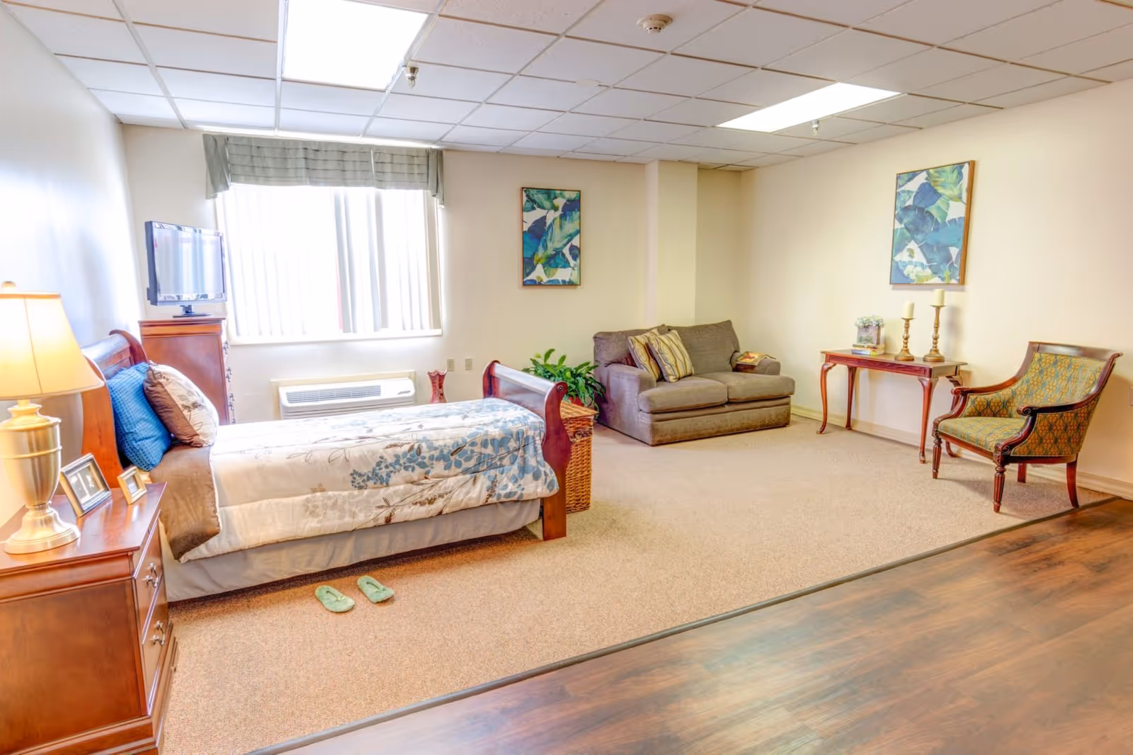 A bright and spacious bedroom in a senior living facility featuring a single bed with floral bedding, a wooden nightstand with a lamp and picture frames, a small flat-screen TV on a dresser, a gray loveseat with striped pillows, a wooden side table with candles and a small plant, an upholstered armchair, and two framed botanical prints on the walls. The room has a large window with vertical blinds and a valance, beige carpet, and wood flooring near the entrance.