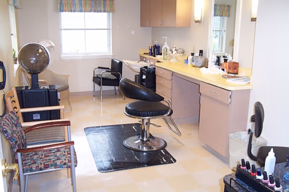 Interior of a salon area in an assisted living facility with a black salon chair on a black mat, a hair dryer, multiple chairs, a sink, and a countertop with various hair care products and supplies.