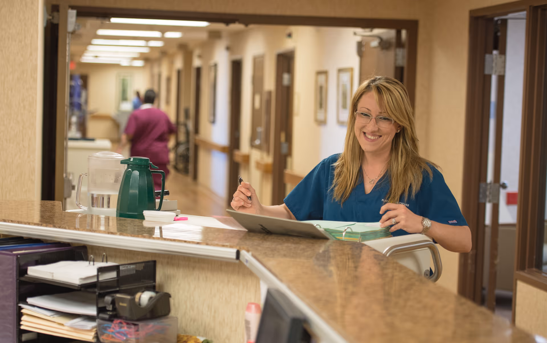 A smiling healthcare worker in blue scrubs stands behind a reception desk in a hallway of a healthcare facility. The desk has a water pitcher, a green thermos, and some papers. Another person in purple scrubs walks down the hallway in the background.