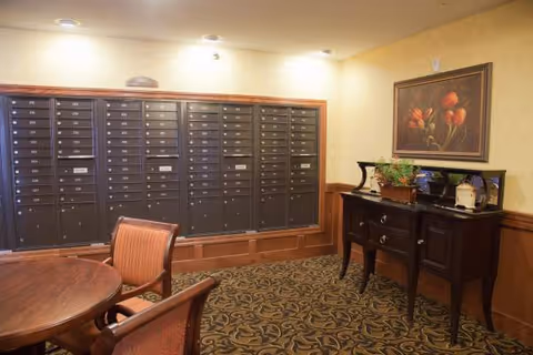 Interior room with a wall of mailboxes, a round wooden table with chairs, a dark wooden side table with decorative items, and a framed painting of flowers on the wall.