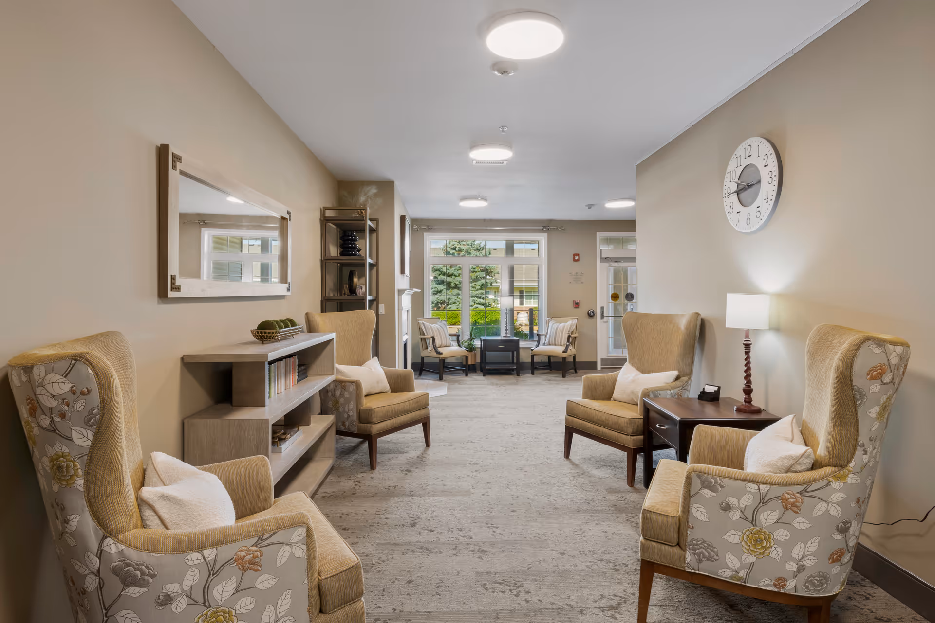 A bright and cozy senior living common area with four upholstered armchairs arranged around a small wooden side table with a lamp. The walls are painted beige, and a large round clock hangs on one wall. A rectangular mirror and a bookshelf with decorative items and books are on the opposite wall. Large windows and a glass door at the far end let in natural light and offer a view of greenery outside.