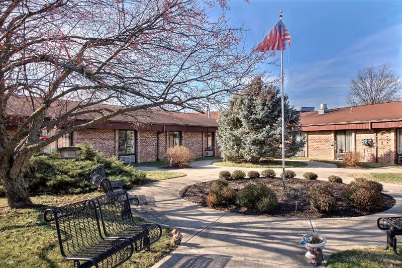 Outdoor courtyard area of a senior living facility with a circular garden bed featuring shrubs and a flagpole with an American flag. Surrounding the courtyard are single-story brick buildings with windows and air conditioning units. There are black metal benches along the paved walkway, and leafless trees indicating a late fall or early winter season.
