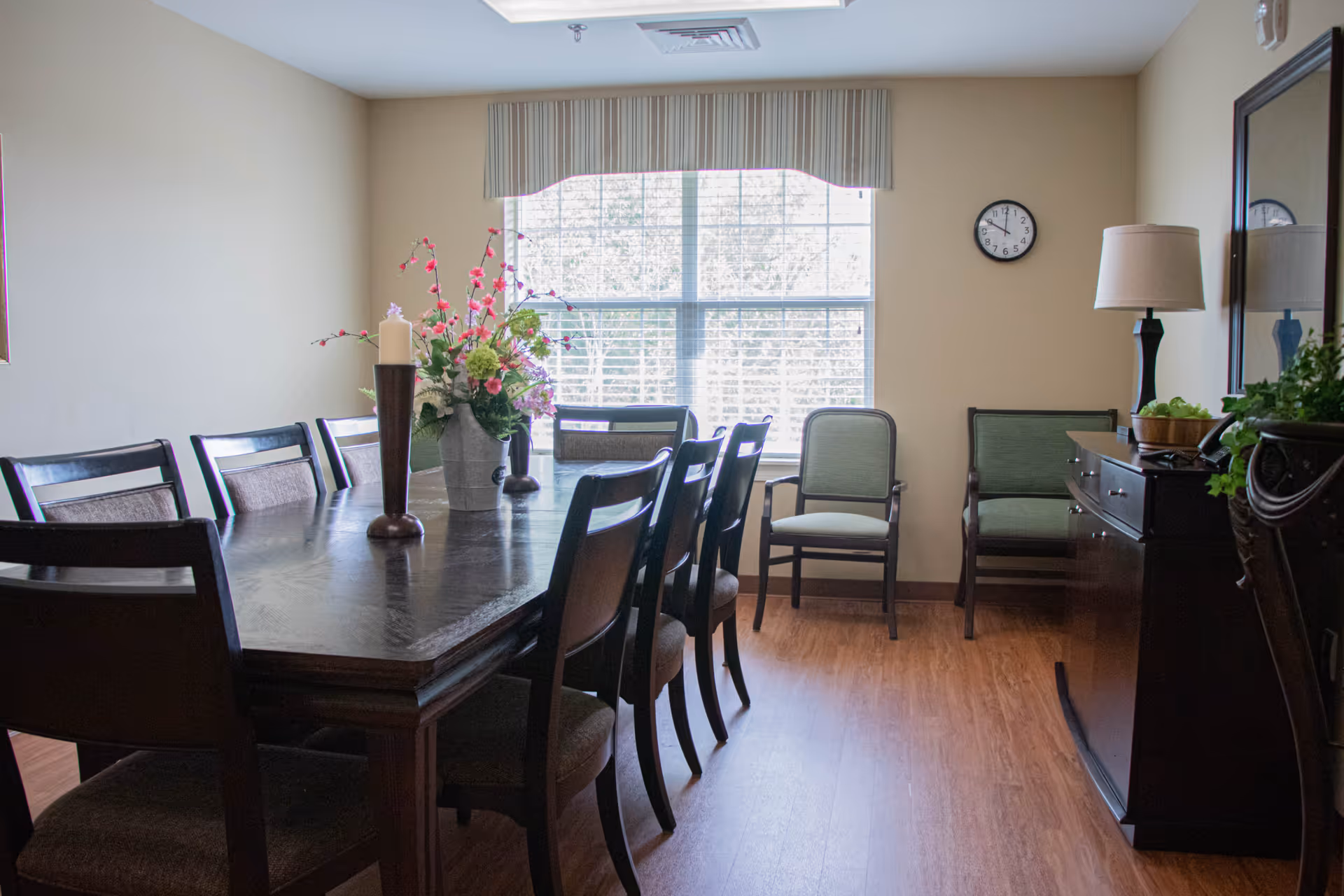 A dining room with a large wooden table surrounded by eight chairs. On the table, there is a floral arrangement and a candle holder. The room has a large window with blinds, two additional chairs against the wall, a clock, a lamp on a sideboard, and a mirror above the sideboard. The floor is wooden.