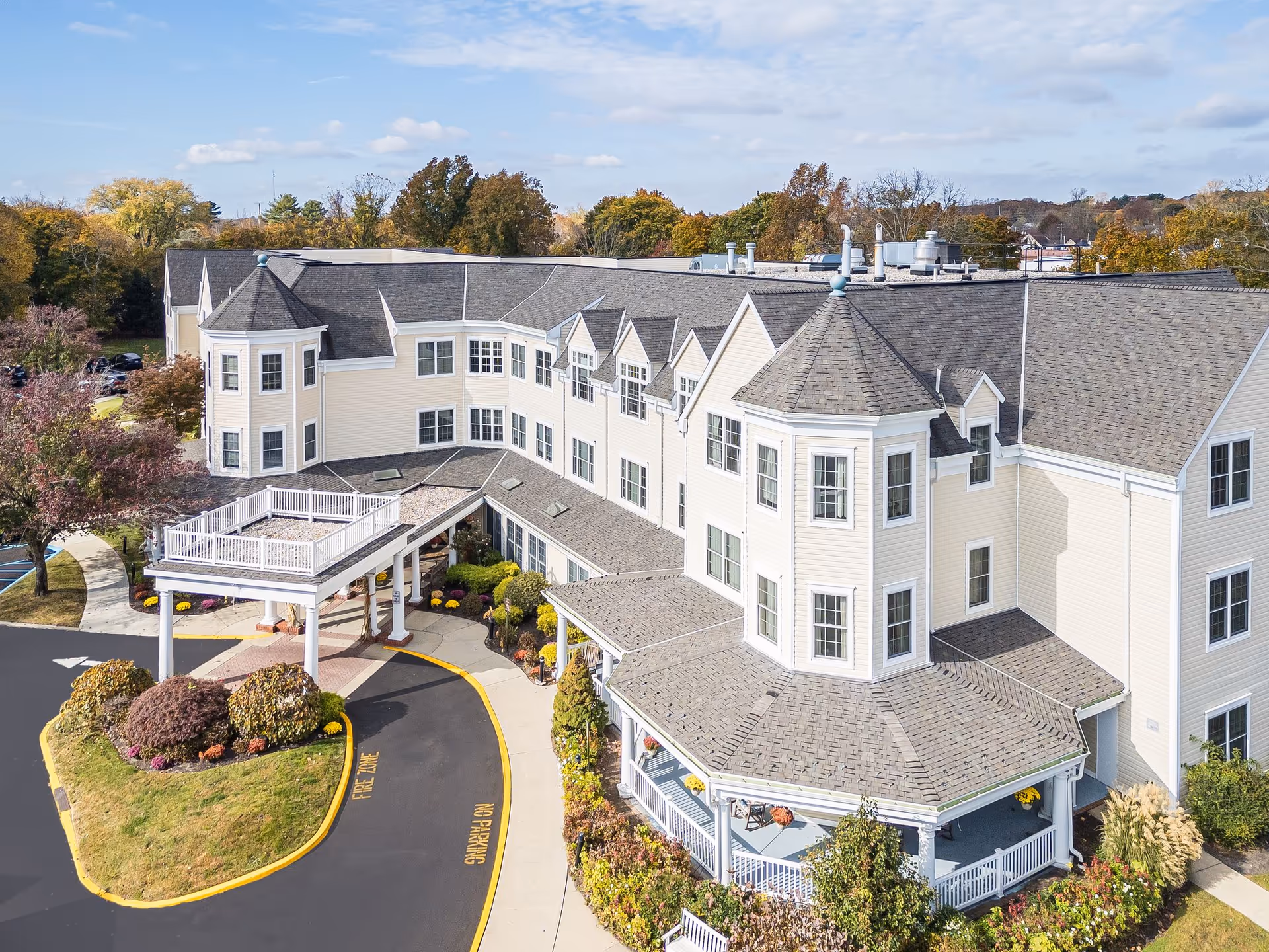 Aerial view of a large, light-colored senior living facility building with multiple windows and a covered entrance driveway. The building has a distinctive architectural design with turret-like structures and a wrap-around porch. Surrounding the building are landscaped gardens with bushes and trees, and a paved driveway marked with 'FIRE ZONE' and 'NO PARKING'. The sky is partly cloudy.
