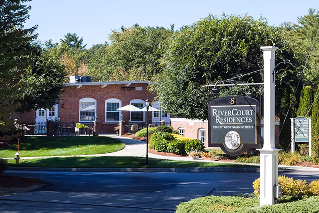 View of the exterior of RiverCourt Residences, showing a brick building surrounded by trees and greenery. A black and white sign with the facility name and address is prominently displayed near the street.