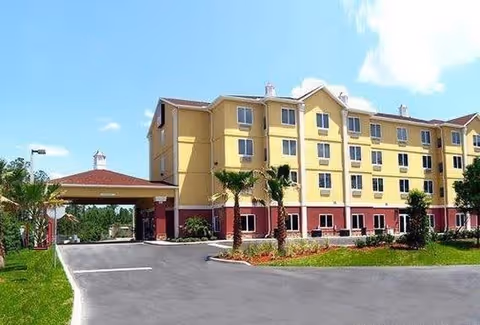 Exterior view of a four-story senior living facility building with a covered entrance driveway, palm trees, and landscaped greenery under a partly cloudy blue sky.