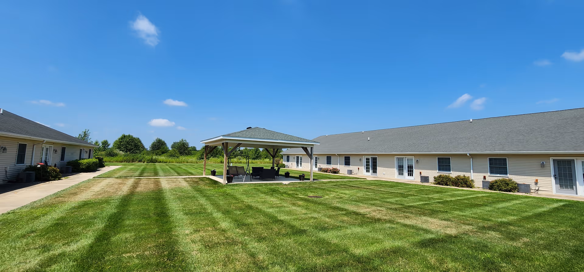 A spacious outdoor courtyard area at The Glenwood Assisted Living of Mahomet featuring a well-maintained green lawn with striped mowing patterns, a covered gazebo with seating, and single-story beige buildings with white doors and windows surrounding the courtyard under a clear blue sky.