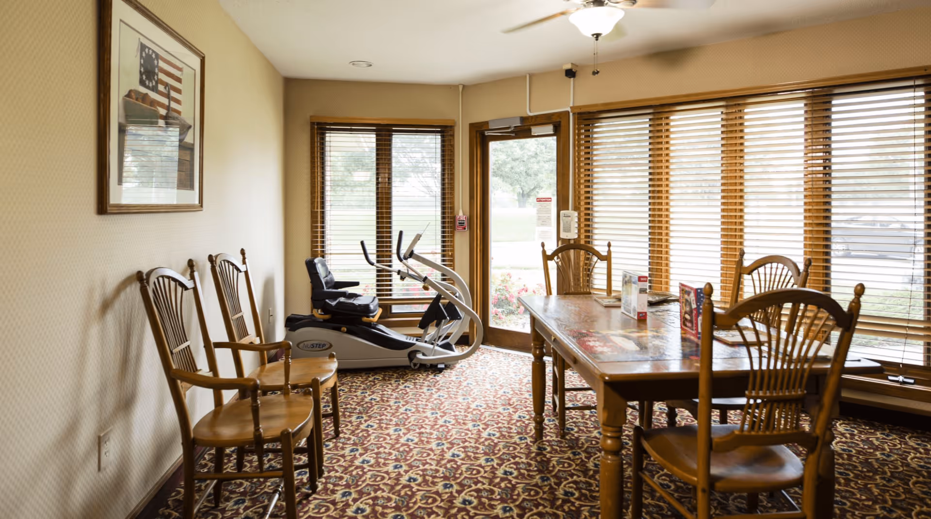 A bright communal room with a wooden dining table and chairs, an exercise machine, and large windows with blinds.