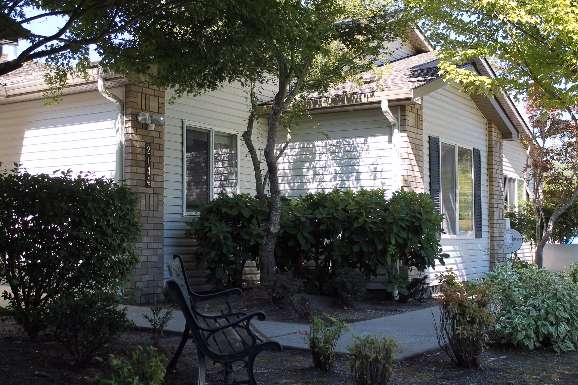 Single-story white siding building front with shrubs, a tree, and benches along a paved walkway.