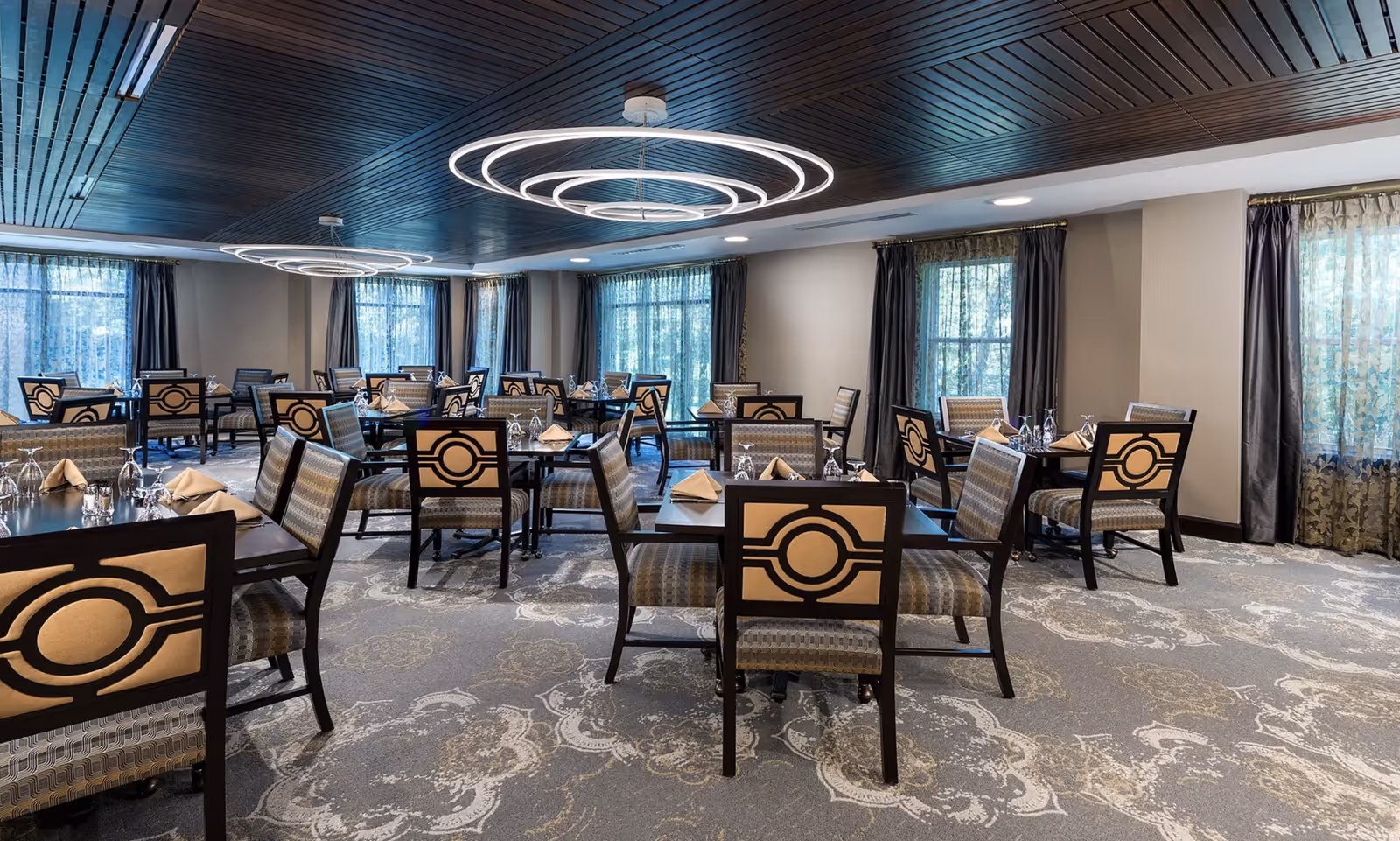 A spacious dining room with multiple dark wood tables and cushioned chairs featuring a circular design on the backrests. The room has large windows with sheer and dark curtains, patterned carpet flooring, and modern circular ceiling lights. The ceiling is dark wood paneled.