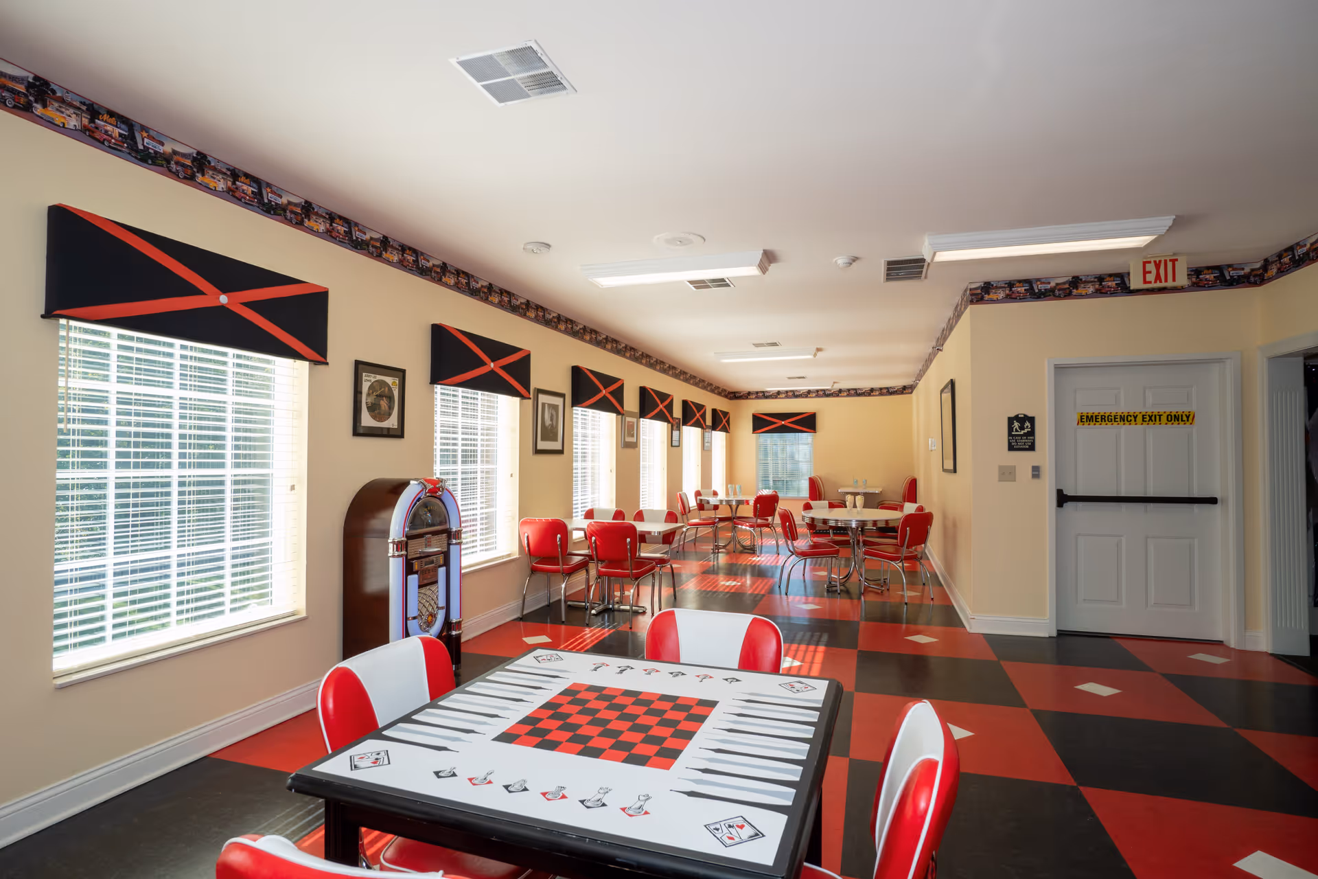 A bright and spacious dining area with red and black checkered flooring and matching red chairs around tables. One table in the foreground has a game board design on its surface. The room has several windows with black and red valances, framed pictures on the walls, and a vintage jukebox near the windows. An emergency exit door is visible at the far end of the room.