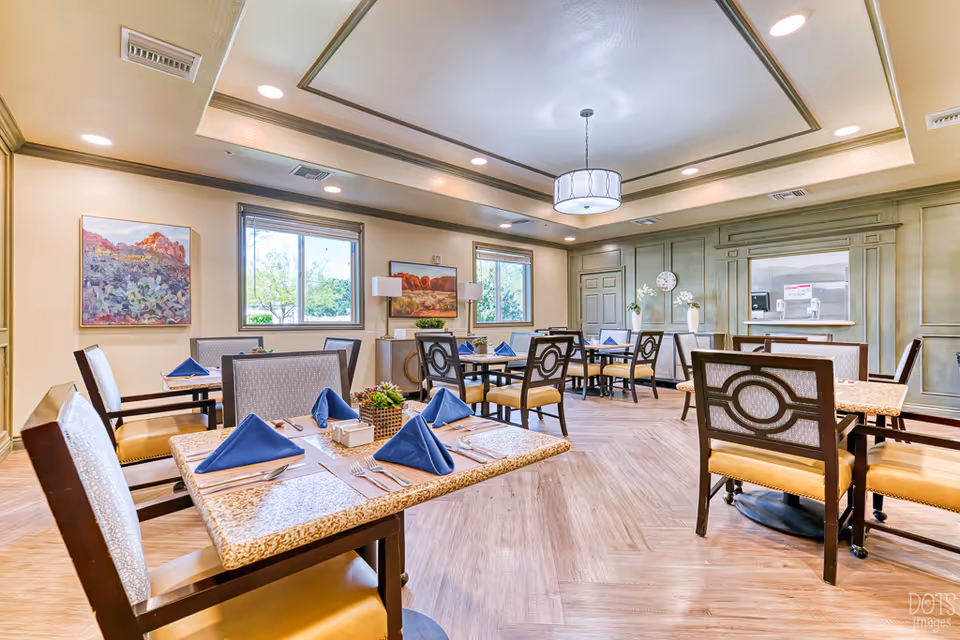 A bright and spacious dining room with several tables set with blue folded napkins, silverware, and small centerpieces. The room features large windows, framed landscape paintings on the walls, a decorative ceiling light fixture, and wood-patterned flooring. The walls have a greenish panel design with a clock and two white vases with flowers on a sideboard.
