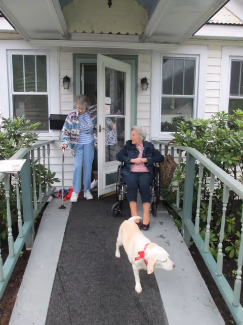 Two elderly women at the entrance of a house; one woman is standing with a cane while the other is sitting in a wheelchair. A yellow dog with a red bow is walking on the ramp leading to the door. The entrance has green railings and plants on both sides.