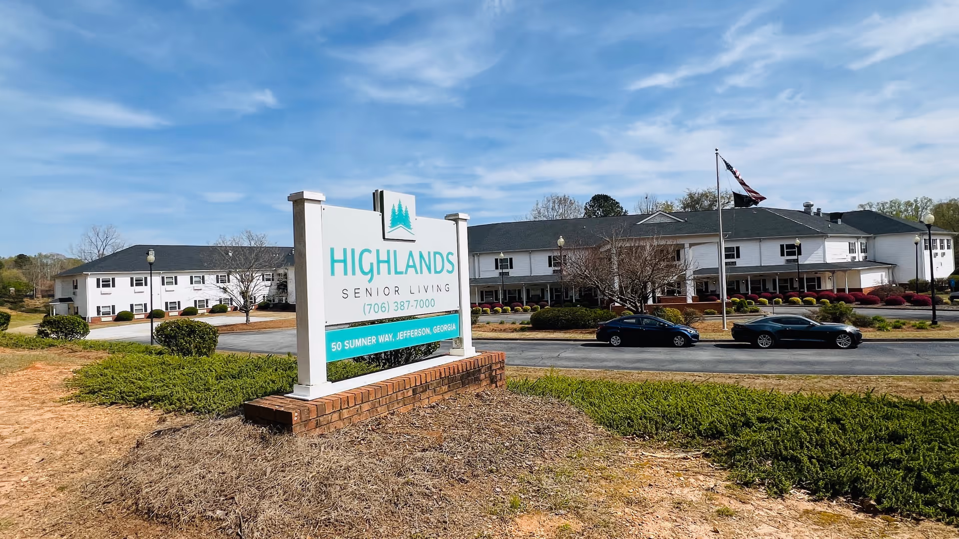 Exterior view of Highlands Senior Living Jefferson facility with a large sign in the foreground displaying the facility name, phone number, and address. The building is a two-story white structure with a dark roof, surrounded by landscaping and a parking area with two cars. A flagpole with a flag is visible near the entrance.