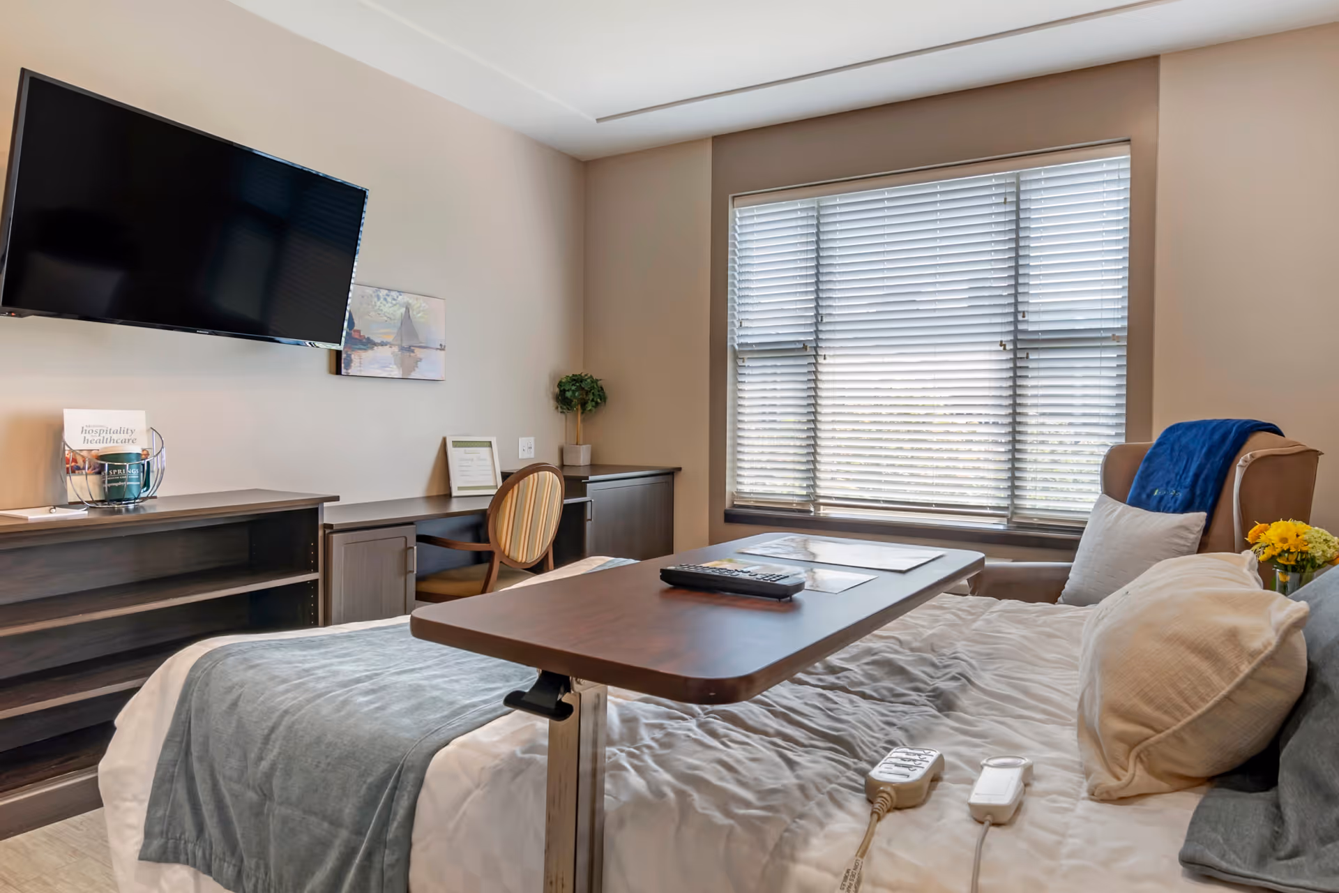 A senior living facility bedroom with a bed covered in white and gray bedding, a wooden overbed table with a remote control on it, a beige armchair with a blue blanket and white pillow, a window with blinds, a wall-mounted flat-screen TV, a desk with a chair, and a small plant on the desk.